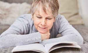 older adult laying on their stomach on a bed, holding open a book with elbows so they can read it